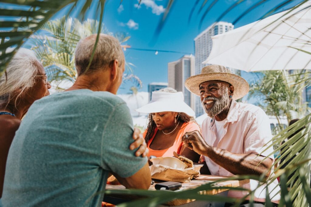 A vibrant African American couple enjoy eating local street food purchased at a farmer's market food fair while dining outdoors with friends they are visiting in Hawaii.