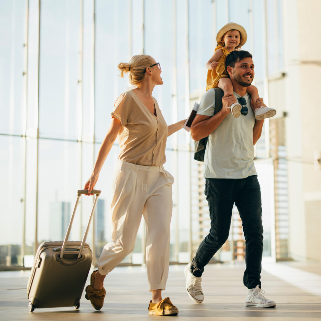 Cheerful husband carrying his cute daughter on shoulders while walking with his beautiful wife and luggage at the airport.