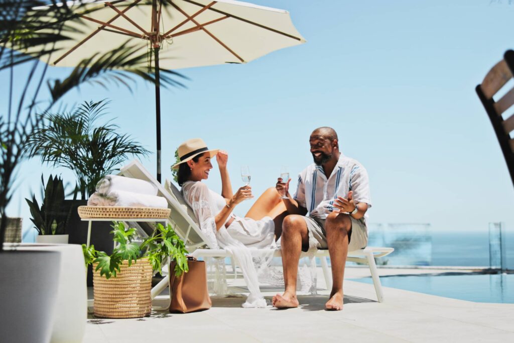 Couple, relax and champagne on vacation on patio, smile or conversation by ocean, celebration or valentines day. Mature people, man and woman with interracial love, drinks or holiday by sea in summer.